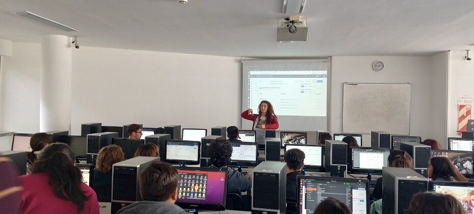 Proyecto de extensión Ciencia de Datos en la escuela: aula con computadoras en la Facultad de Informática. De frente la profesora hablando, detrás una pantalla con una presentación. Estudiantes de espaldas mirando a la profesora.