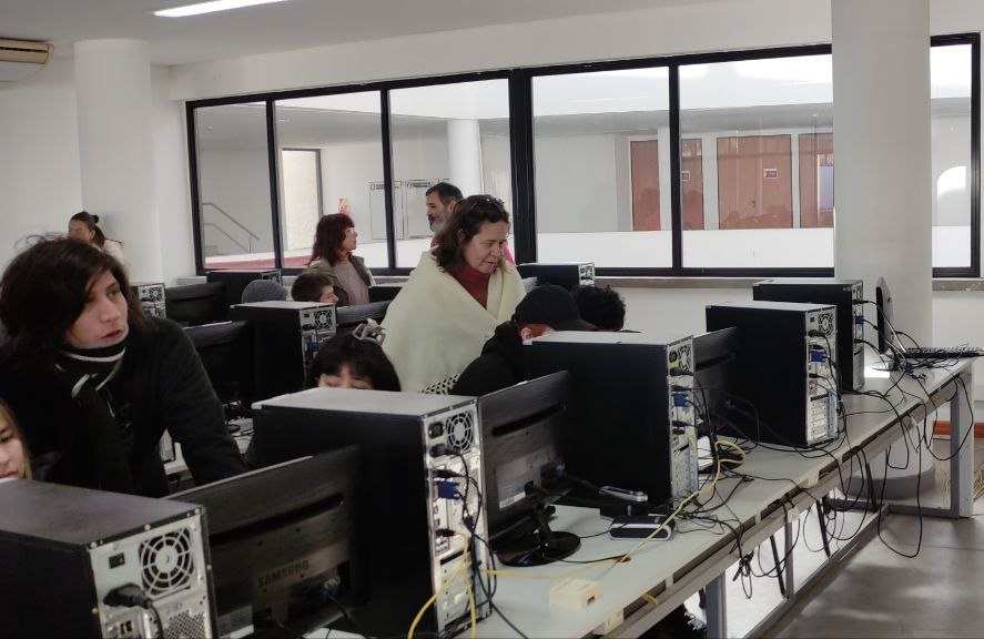 Sala de PC de la Facultad de Informática de la UNLP con docentes y estudiantes, trabajando en computadoras de escritorio. Se observan filas de equipos conectados y personas interactuando en un ambiente luminoso con grandes ventanas. La escena refleja una actividad educativa en un espacio moderno.