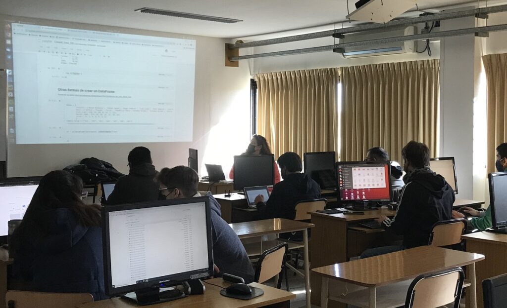 Aula de la Facultad de Informática de la UNLP con docentes y estudiantes, trabajando en computadoras de escritorio. Se observa la proyección de una presentación y personas interactuando en un ambiente semioscuro. La escena refleja una actividad educativa en un espacio moderno.
