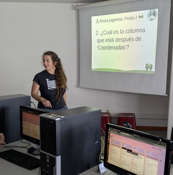 Aula de la Facultad de Informática de la UNLP con una docente y estudiantes, trabajando en computadoras de escritorio. Se observa la proyección de una presentación y personas interactuando en un ambiente semioscuro. La escena refleja una actividad educativa en un espacio moderno.