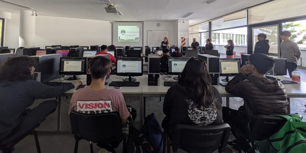 La imagen muestra un aula de informática con varias personas, principalmente estudiantes, sentadas frente a computadoras de escritorio distribuidas en filas. Al fondo, un grupo de docentes o facilitadores está de pie, aparentemente impartiendo una clase o presentación, mientras en la pantalla del proyector se observa contenido relacionado con la lección. La habitación es amplia y luminosa, con grandes ventanas a la derecha que permiten la entrada de luz natural. Los estudiantes están atentos a sus pantallas o a la explicación dada, reflejando un ambiente de aprendizaje tecnológico y colaborativo.