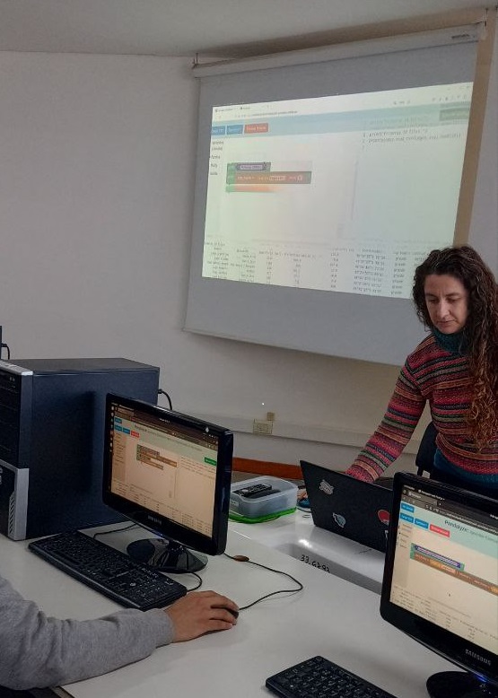 Aula de la Facultad de Informática de la UNLP con una docente y estudiantes, trabajando en computadoras de escritorio. Se observa la proyección de una presentación y personas interactuando en un ambiente semioscuro. La escena refleja una actividad educativa en un espacio moderno.