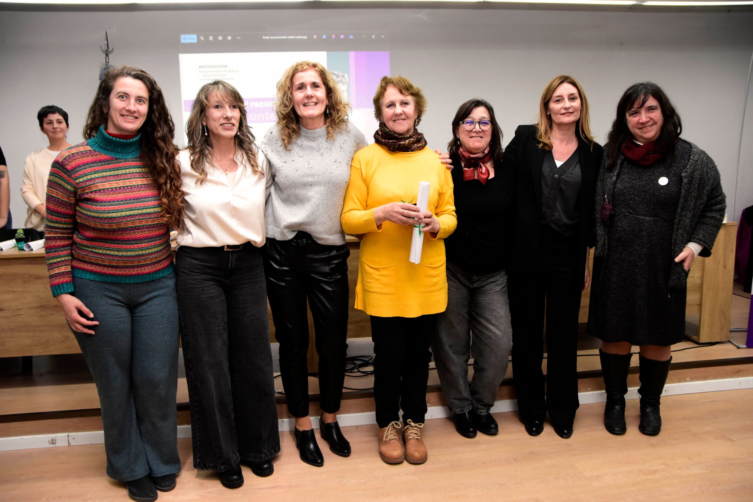 Andrea Varela, Verónica Cruz, Ana Barletta, Lía Molinari, Ivana Harari, Sofía Martin y Claudia Queiruga posando juntas en un salón interior. Están de pie, vestidas de manera casual y semiformal, en un salón con piso de madera y una pantalla proyectada al fondo. Lía Molinari en el centro, con suéter amarillo, sostiene el diploma enrollado del Reconocimiento "Julieta Lanteri" otorgado por la UNLP. La imagen transmite un ambiente de reconocimiento y celebración.