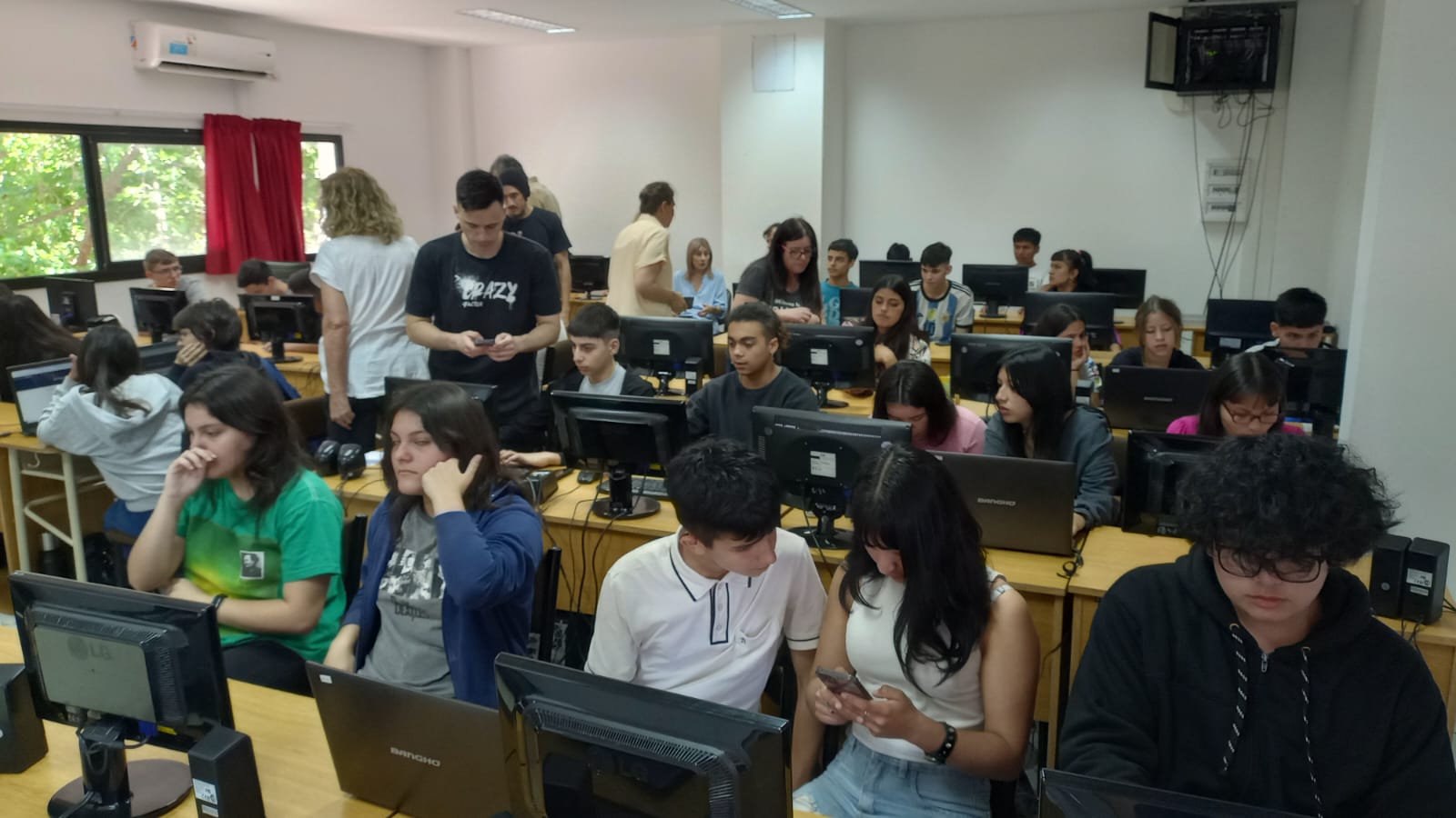 Salón de clases lleno de estudiantes frente a computadoras en una galería de dos filas. Varias personas trabajan en laptops, algunas conversan y otros miran sus pantallas. El ambiente es de aprendizaje colaborativo, con docentes y asistentes de fondo apoyando. Se observa una mezcla de chicos y chicas de diferentes edades, ropa casual y un ambiente tecnológico con múltiples monitores y equipos informáticos.