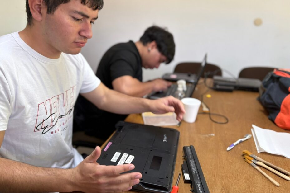 En un aula de la Facultad de Informática en el marco de las Prácticas Profesionalizantes Supervisadas se ve en primer plano, un estudiante secundario con camiseta blanca que sostiene una notebook volteada, mientras otra persona al fondo trabaja con su equipo. En la mesa hay herramientas y una funda de laptop negra. Ambiente de aula o taller.